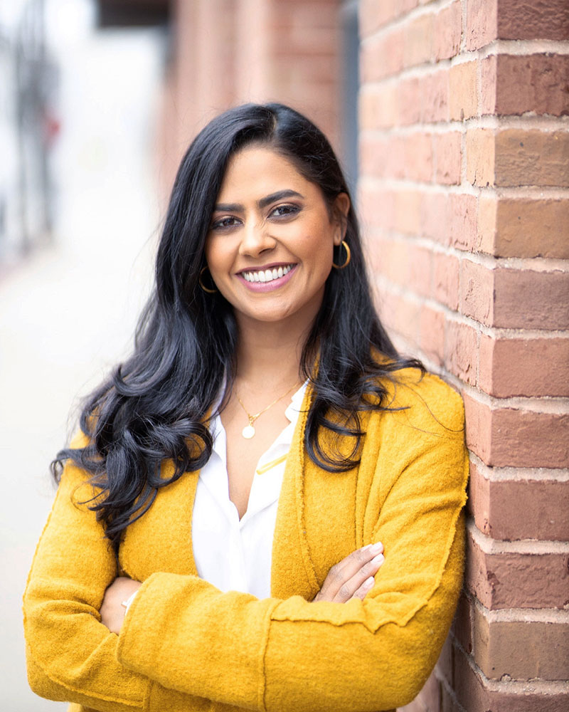 Dr. Veena Vaidyanathan smiles while leaning on a building wall.